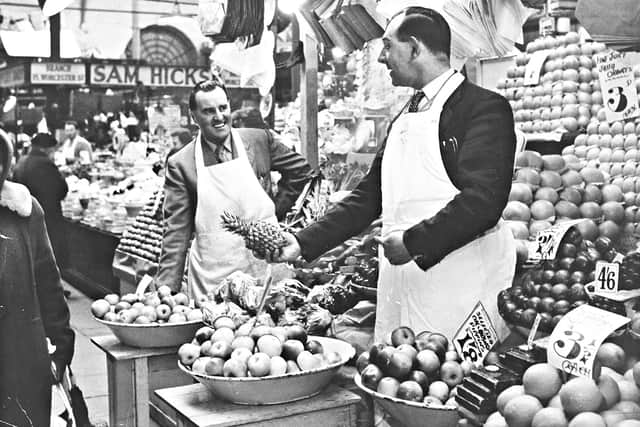 Ronald Gough (right) and his cousin Jim standing at the front of their fruit stall at Old Retail Market Hall, North Street, May 1960. Ron is holding out a pineapple to Jim. There is a sign for "Sam Hicks" visible in the background.