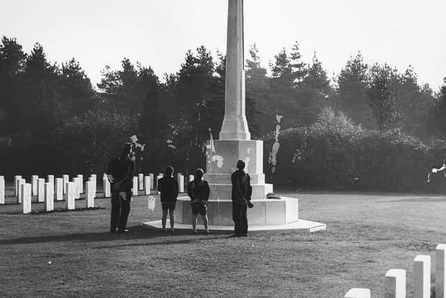 Cross of Sacrifice, Cannock Chase Military Cemetery. This photograph shows the Cross of Sacrifice, surrounded by headstones, with a man and three boys standing near the cross and looking up at it on November 8, 1959. A wreath is visible on the base of the cross. The cemetery is maintained by the Commonwealth War Graves Commission (CWGC)