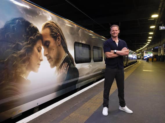 Sam Heughan in front of a train with an Outlander wrap