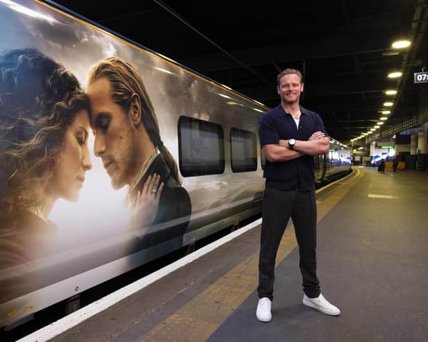 Sam Heughan in front of a train with an Outlander wrap