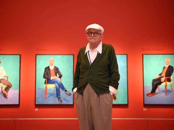David Hockney with some of his portraits behind him hung on a red wall