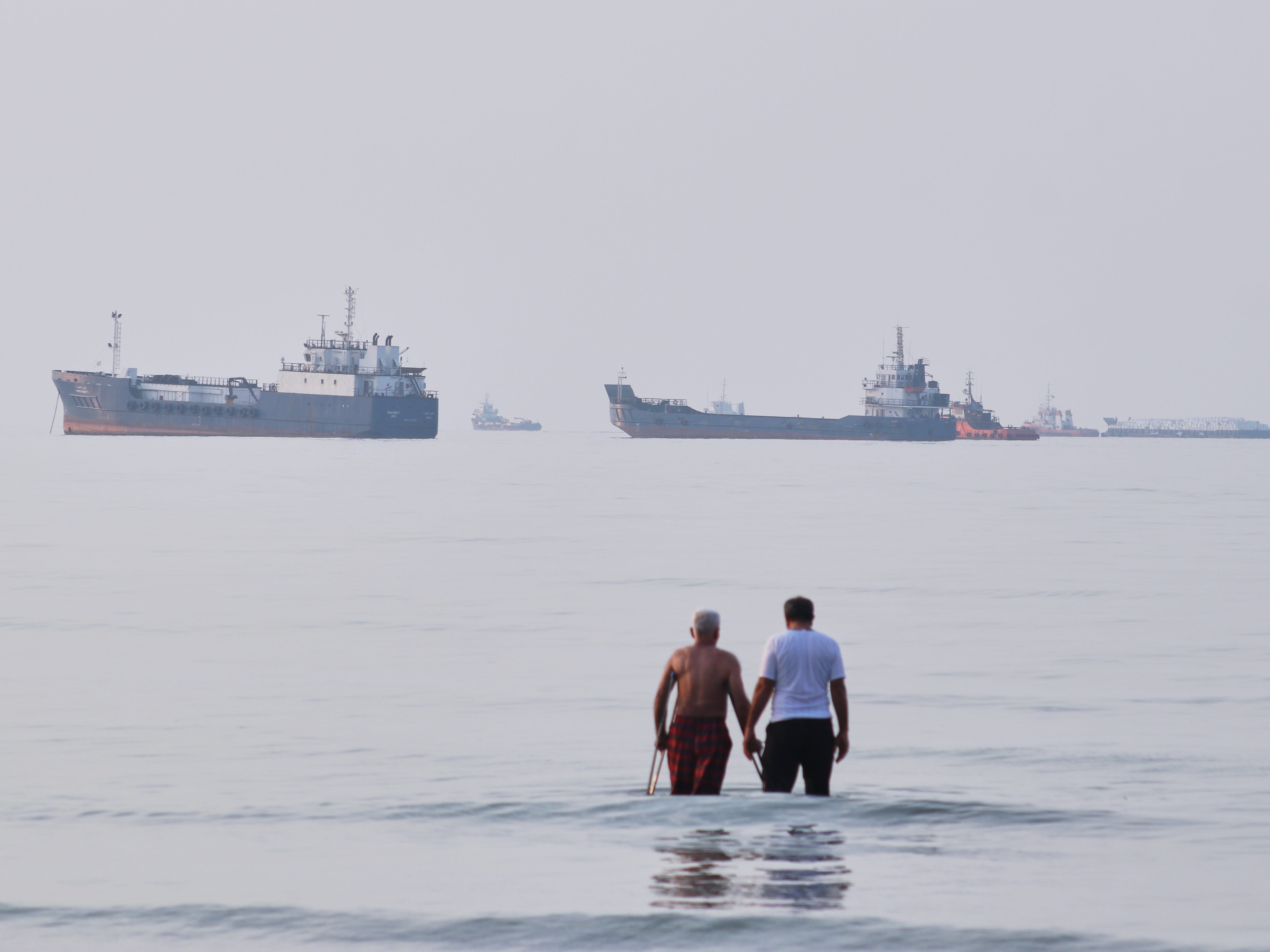 Two men wade in the waters of the Strait of Hormuz with vessels anchored in the background (Razieh Poudat/AP)
