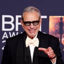 Jeff Goldblum smiling with his left hand raised and pointing wearing a black suit and white bow tie at the Brit Awards