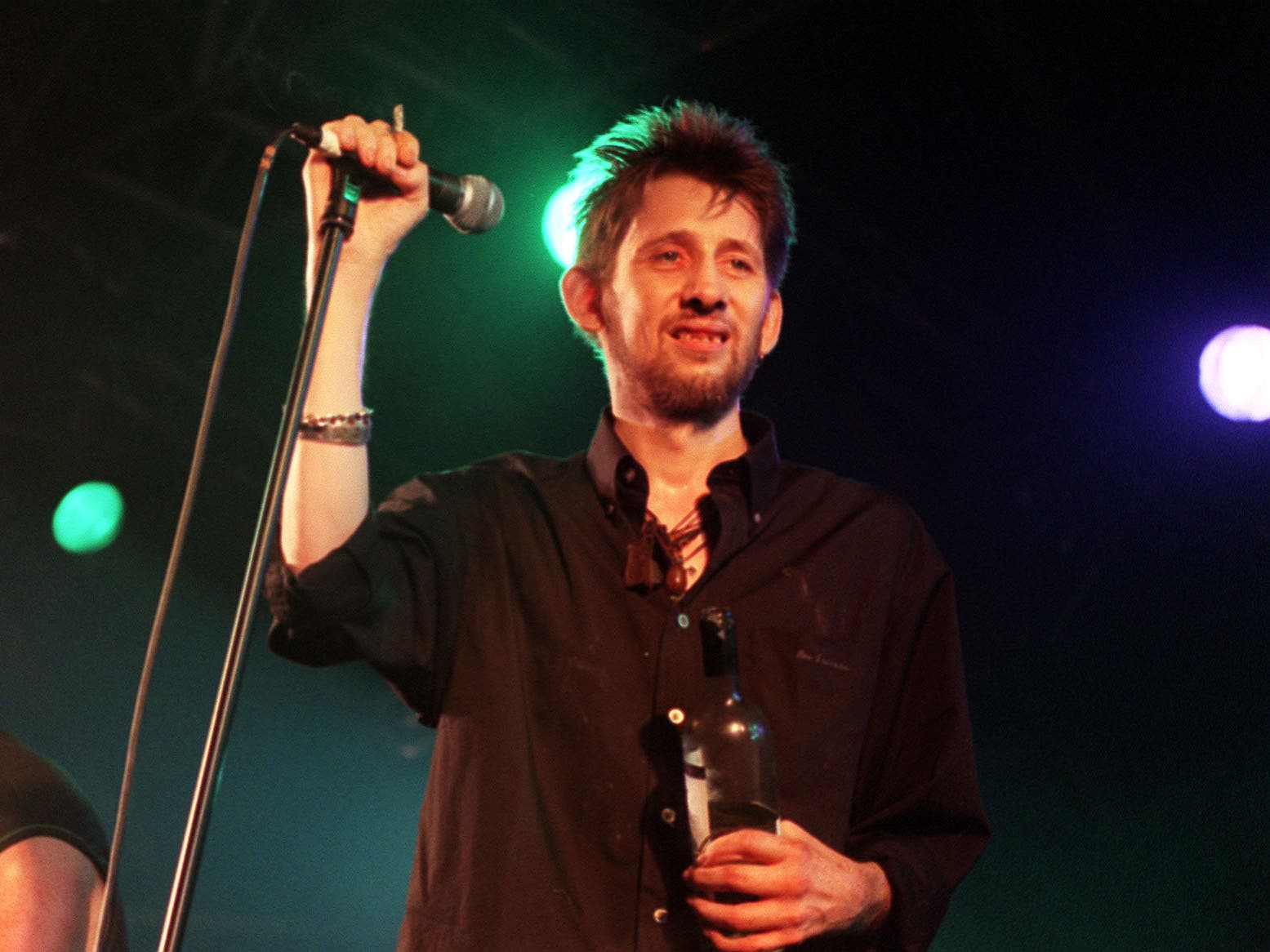 Shane MacGowan smiling while on stage, holding a microphone stand