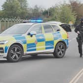 An armed policeman walking on a road, next to a police car with its blue lights on