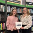 Lola (left) and Cherry (right) stand in front of bookshelves in Stafford Library, holding a Staffordshire Poet Laureate folder beneath the library's fiction section signage.