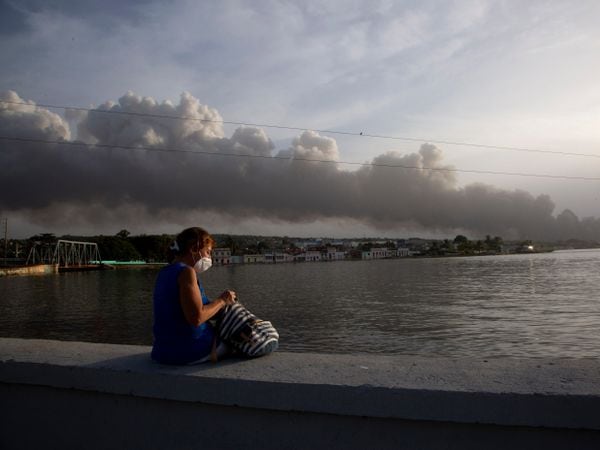 A resident sits on the sea wall as smoke rises in the background from a deadly fire at a large oil storage facility in Matanzas, Cuba