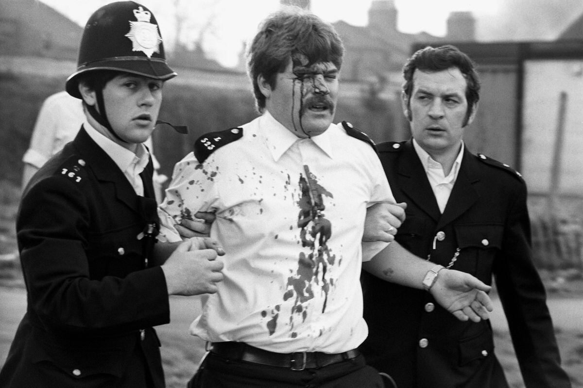 A police officer, blood streaming from a head wound, being helped away by colleagues during rioting in Brixton